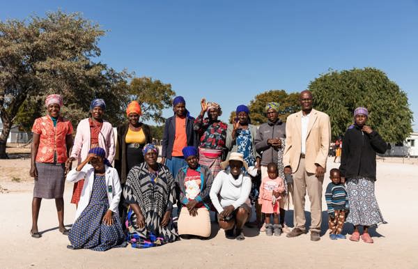 A large group of men, women, and children in a mix of modern and traditional clothing pose for a group photo outdoors in a sunny Botswanan village setting.