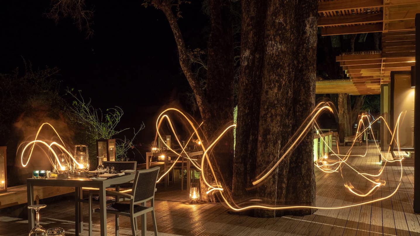 Long exposure 'light painting' photography of an outdoor dining area at Vumbura Camp, with light trails weaving around tree trunks.