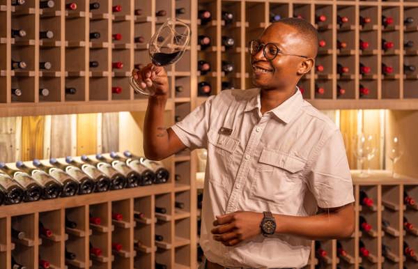 A smiling safari lodge sommelier or staff member inspecting a glass of red wine inside the exclusive Mombo Camp wine cellar, with bottles stored in modern wooden racking, highlighting the gourmet experience.