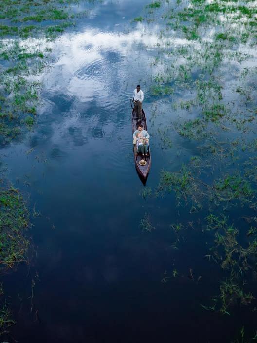 High overhead aerial view of a poler guiding a mokoro canoe with tourists through the dark, reflective, and grass-filled waters of the Okavango Delta, Botswana.