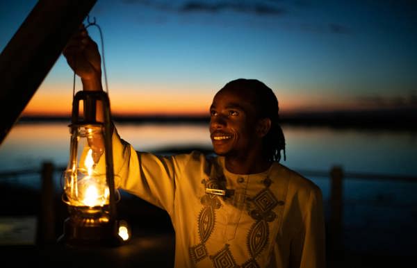 A friendly safari lodge staff member in Botswana, dressed in a traditional patterned shirt, smiles while holding a glowing lantern against a vivid sunset over the water, highlighting the exclusive and personal service.