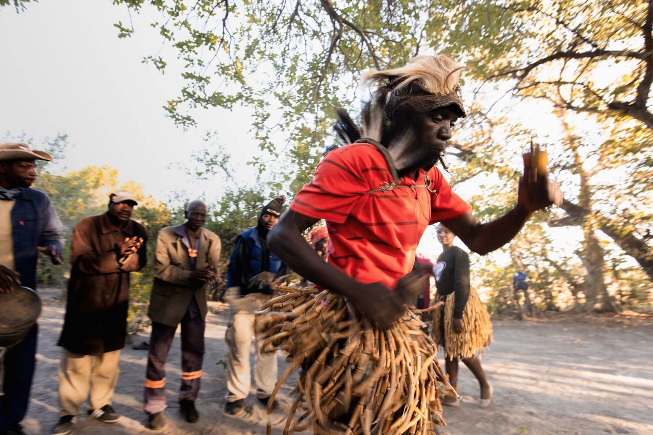 A man performing a traditional Botswana dance in a skirt made of reeds or sticks, with onlookers and drumming in a natural setting.