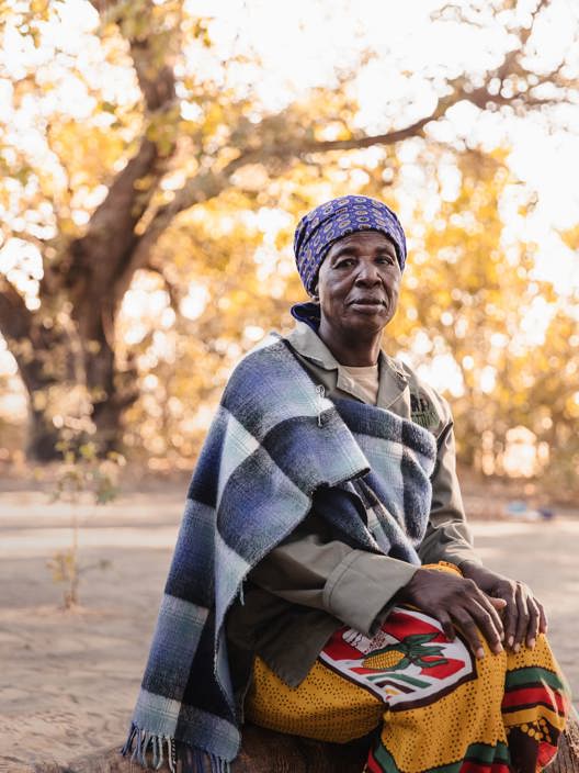 A portrait of an older woman in a blue head wrap, checkered shawl, and colorful patterned skirt, sitting outdoors in a village setting with golden light filtering through the trees.