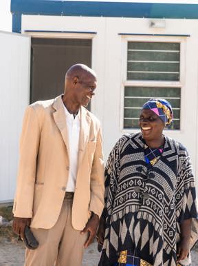 A smiling older man in a light suit and a woman in traditional patterned dress and head wrap stand outside a white portable building in a Botswanan village.