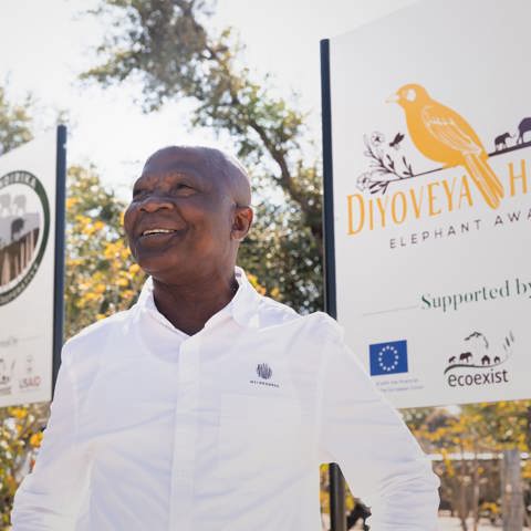 A man smiling in front of a sign for 'Diyoveya Honey, Elephant Aware', highlighting community conservation and honey making in Botswana.