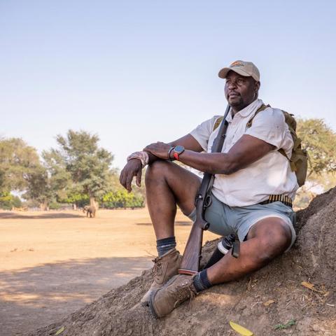 A guide sits on a termite mound, with a backpack and a rifle.