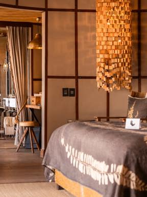 The stunning interior of a luxury safari suite bathroom, featuring a central bronze freestanding bathtub, a marble double vanity, and large open shutters that lead to a private view of the African wilderness.