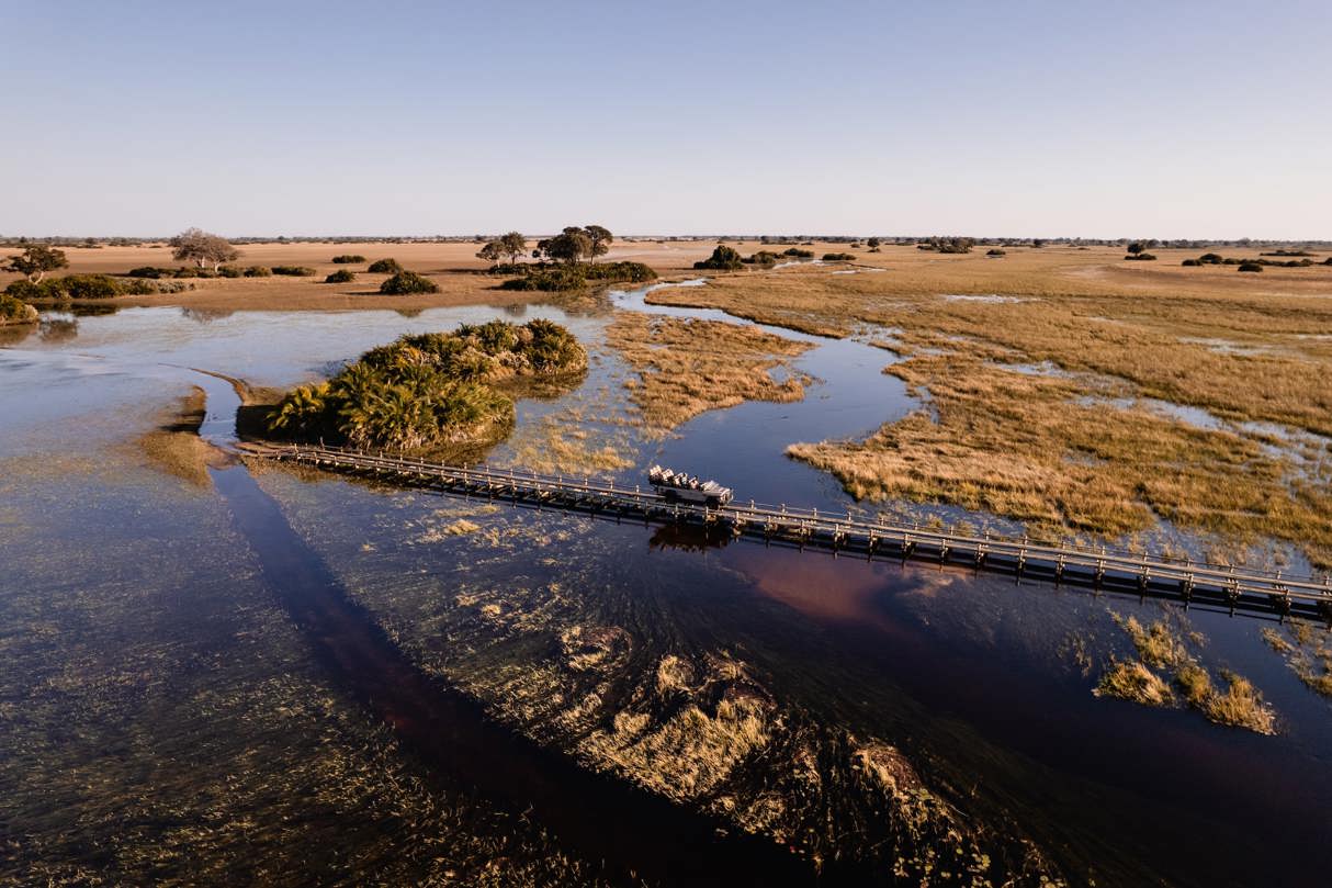 Aerial view of a safari game drive vehicle with tourists crossing a long, narrow wooden bridge over a deep channel in the vast Okavango Delta landscape.