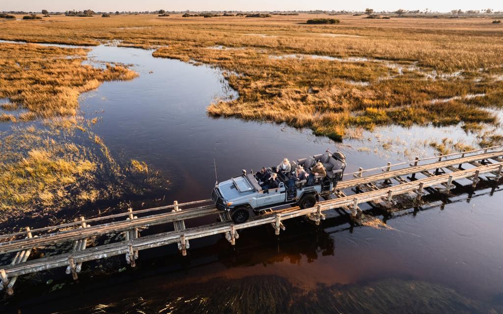 High angle aerial shot of a safari game drive vehicle with tourists crossing a wooden bridge over a water channel in the Okavango Delta, surrounded by golden marshland.