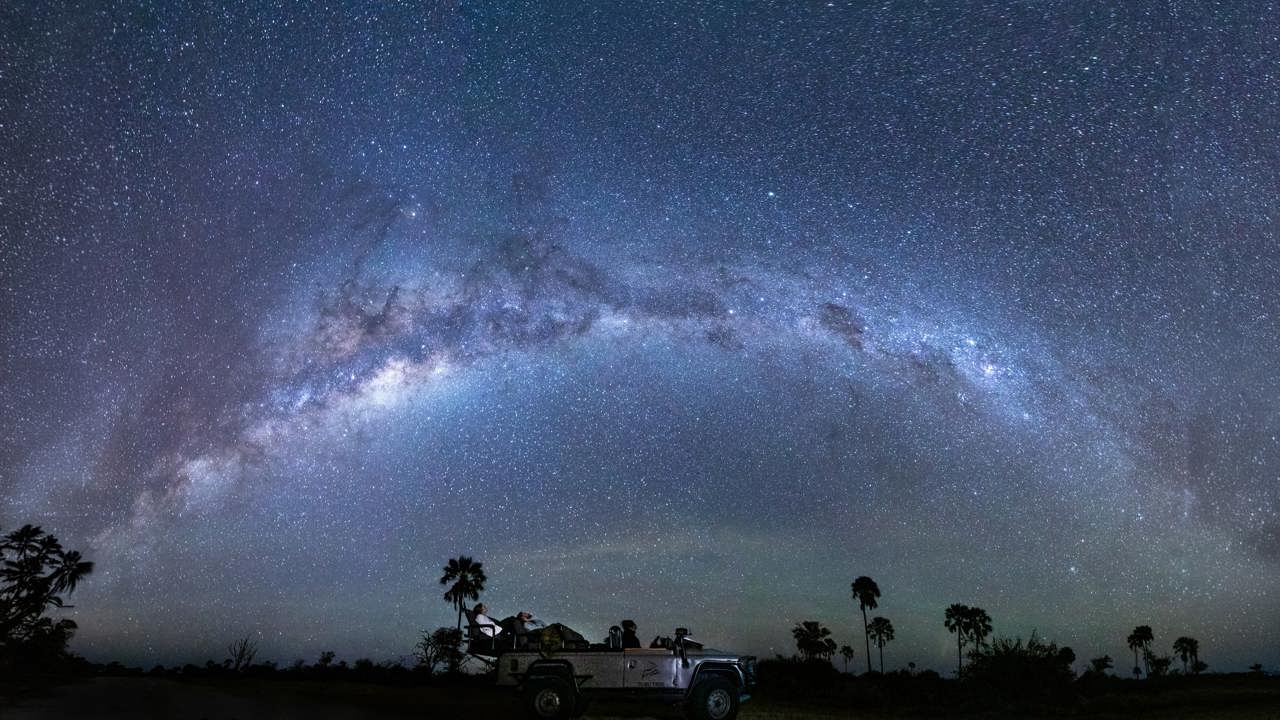 A safari vehicle with tourists parked under a brilliant, sweeping arc of the Milky Way galaxy during a star-gazing safari night drive in Botswana.